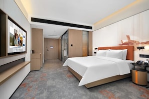 Ein modernes Hotelzimmer mit einem großen Bett, einem an der Wand montierten Fernseher und einem durch eine Glastür einsehbaren Badezimmer.