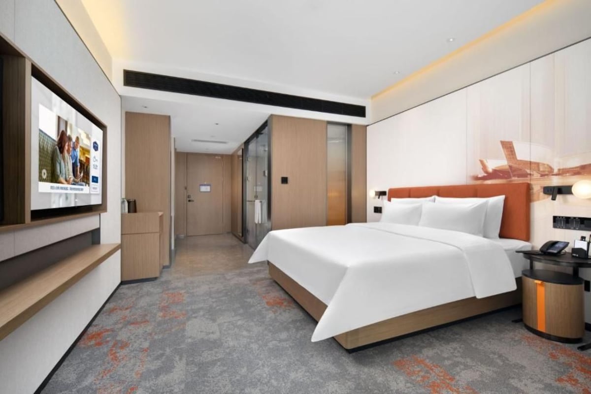 Una habitación de hotel con una cama grande, una mesita de noche con lámpara, un televisor montado en la pared y un cesto de basura pequeño.