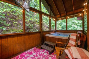 A wooden cabin with a hot tub, a bed with a pink floral blanket, and a view of a forested area. - 2Mi to Pkway|Hot Tub|Newly Renovated|LUXE Cabin (Pigeon Forge)
