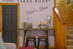 A room with a wooden door, a wall with the phrase 'Love Takes Root,' a small table with books and a vase of flowers, and two stools. - 2Mi to Pkway|Hot Tub|Newly Renovated|LUXE Cabin (Pigeon Forge)