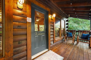 A wooden cabin with a dark front door, a welcome sign, a porch with a rocking chair, and a view of the surrounding forest. - 2Mi to Pkway|Hot Tub|Newly Renovated|LUXE Cabin (Pigeon Forge)