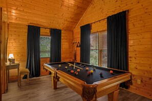 Ein Zimmer aus Holz mit einem Billardtisch, zwei mit Vorhängen versehenen Fenstern, einer Lampe und einem Sessel.