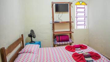 Una camera piccola con un letto singolo, una testiera in legno, una ventola sul comodino, una TV montata su una mensola di legno, una finestra con cornice viola e una coperta colorata.