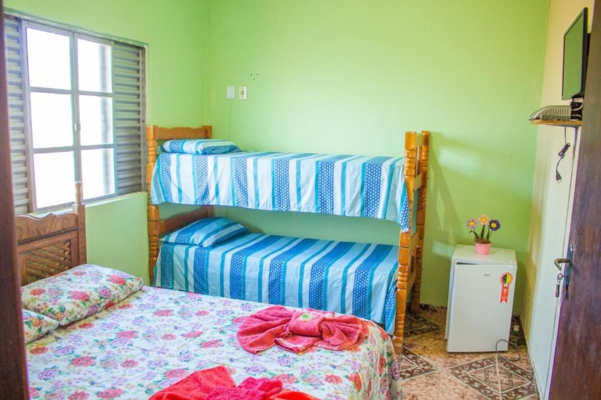 Une chambre avec un lit superposé, un lit simple, un petit réfrigérateur et une fenêtre avec des stores.