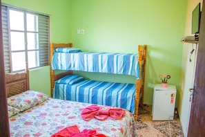 Une chambre avec un lit superposé, un lit simple, un petit réfrigérateur et une fenêtre avec des stores.