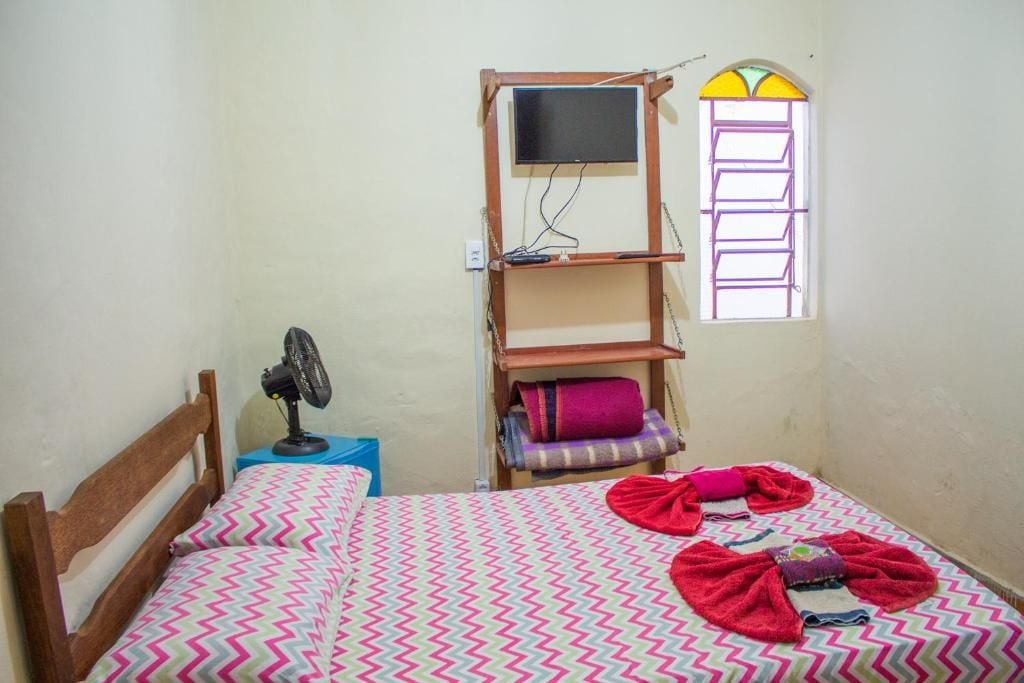 Una habitación pequeña con cama individual, cabecera de madera, ventilador de pie, televisor montado en estante de madera, ventana con marco morado y colcha colorida.