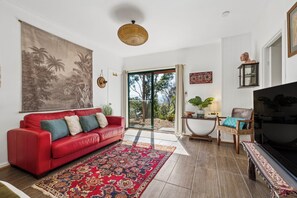 Una sala de estar con un sofá rojo, un tapiz grande en la pared, una lámpara de techo tejida, un televisor y vistas a través de una puerta de cristal.