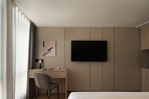 A hotel room with a bed, a desk with a chair, a television mounted on the wall, and a window with curtains.