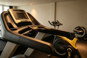 Una bicicleta estaciónia amb pantalla digital i panell de control, situada en una habitació amb una cinta de córrer i equip d'exercici fixat a la paret.