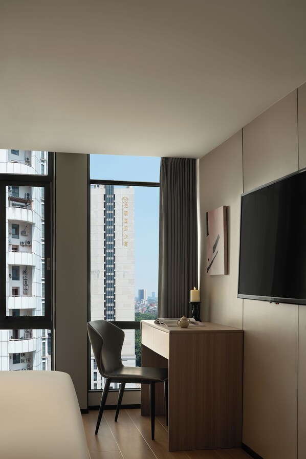 A modern hotel room with a large window, a desk, a chair, a bed, and a flat-screen TV.