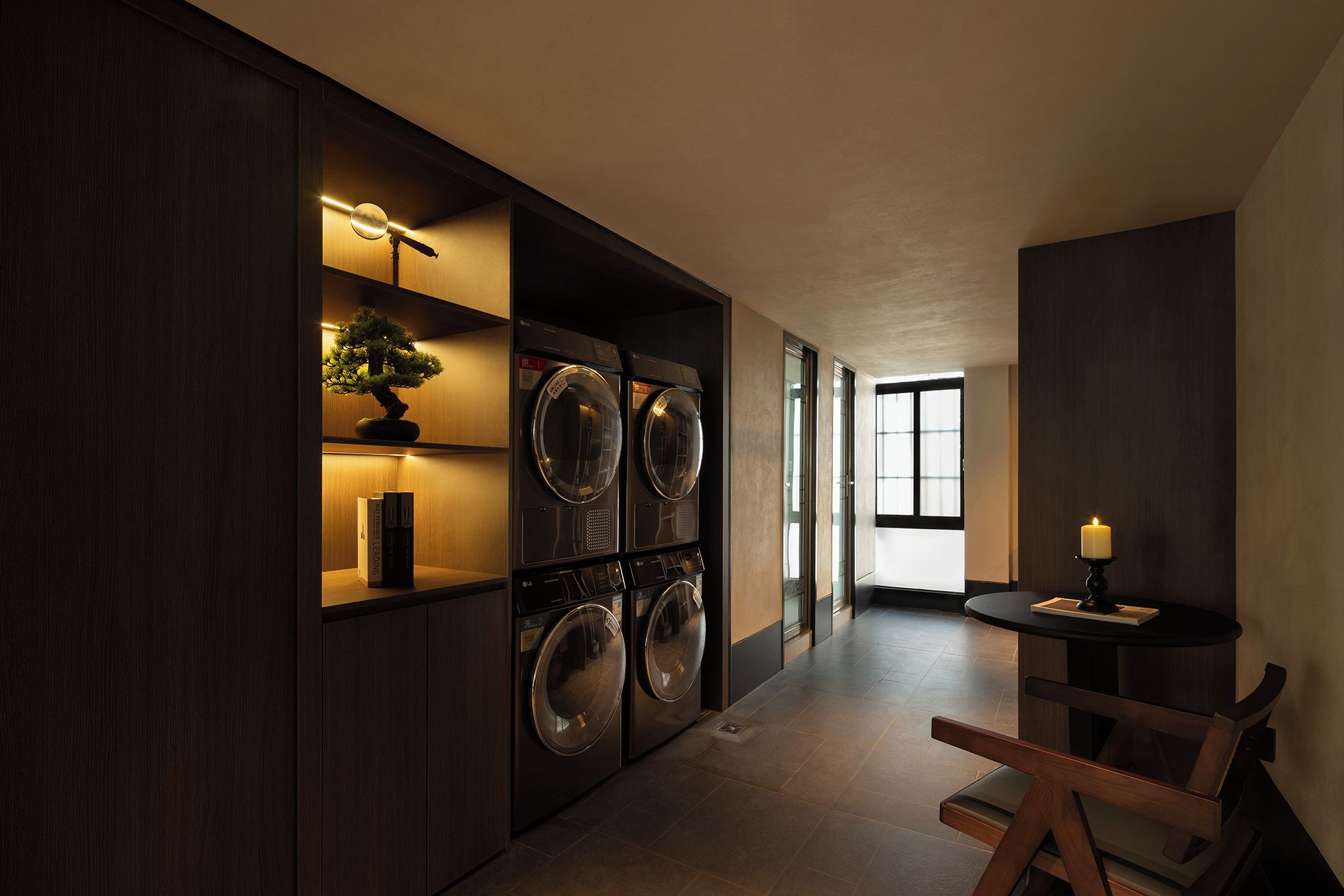 A laundry room with multiple washing machines, a small table with a candle, and a bonsai tree on a shelf.