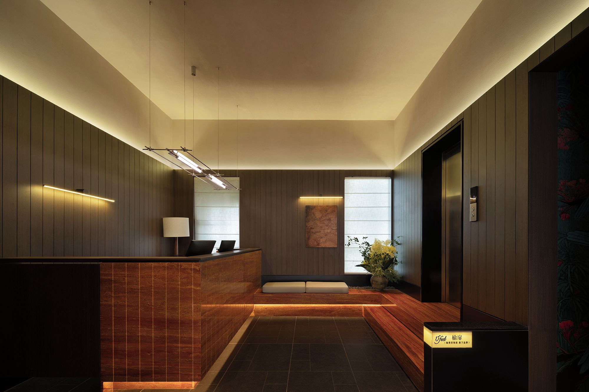 A modern reception area with a wooden reception desk, a seating area with cushions, a large window with natural light, and a wall-mounted artwork.