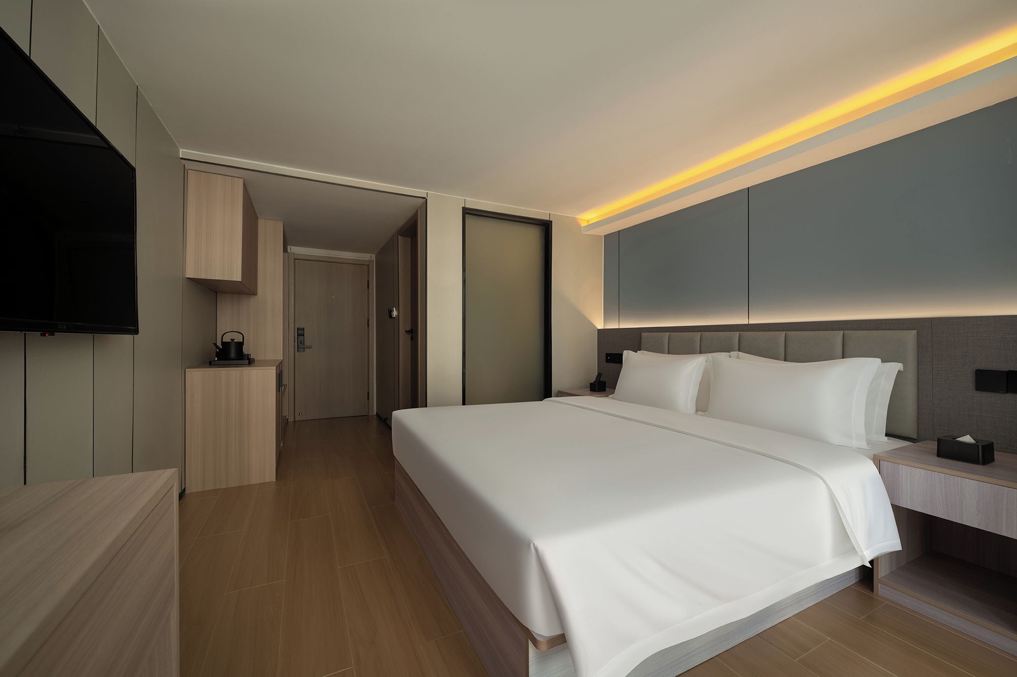 A modern hotel room with a large bed, a flat-screen TV mounted on the wall, and a bedside table with a lamp.