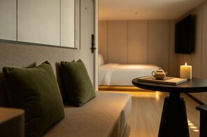 Habitación de hotel con sofá, mesita de noche con tetera y un libro, vela encendida y cama al fondo.