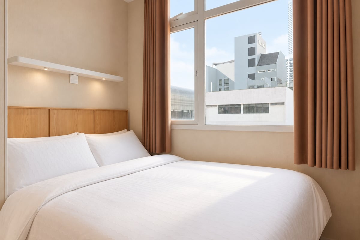 A hotel room with a large bed, wooden headboard, and a view of a cityscape through a window.
