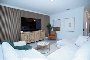 A modern living room with a flat-screen TV, a wooden cabinet, a brown armchair, a white sofa, and two framed artworks on the wall. - 6BR Pool Home w/ Theater & Game Room (Davenport)