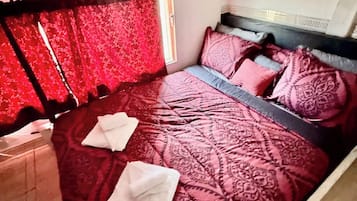 A bedroom with a large bed, a headboard, and a window with red curtains.