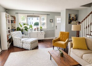 Ein Wohnzimmer mit Sofa, Sessel, Hocker, Couchtisch, Bücherregal und einem Fenster mit Jalousien.