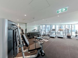 Academia moderna com diversos equipamentos de exercício, janelas amplas e sinalização de saída visível.