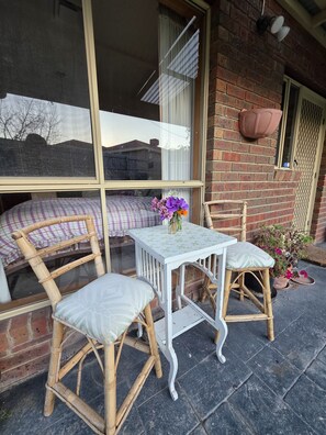 A patio with two wicker chairs, a small white table with a vase of flowers, and a view through a large window. - 🌿 Private Queen Bedroom, Private Bathroom & Garden Retreat in Sunbury (Sunbury)