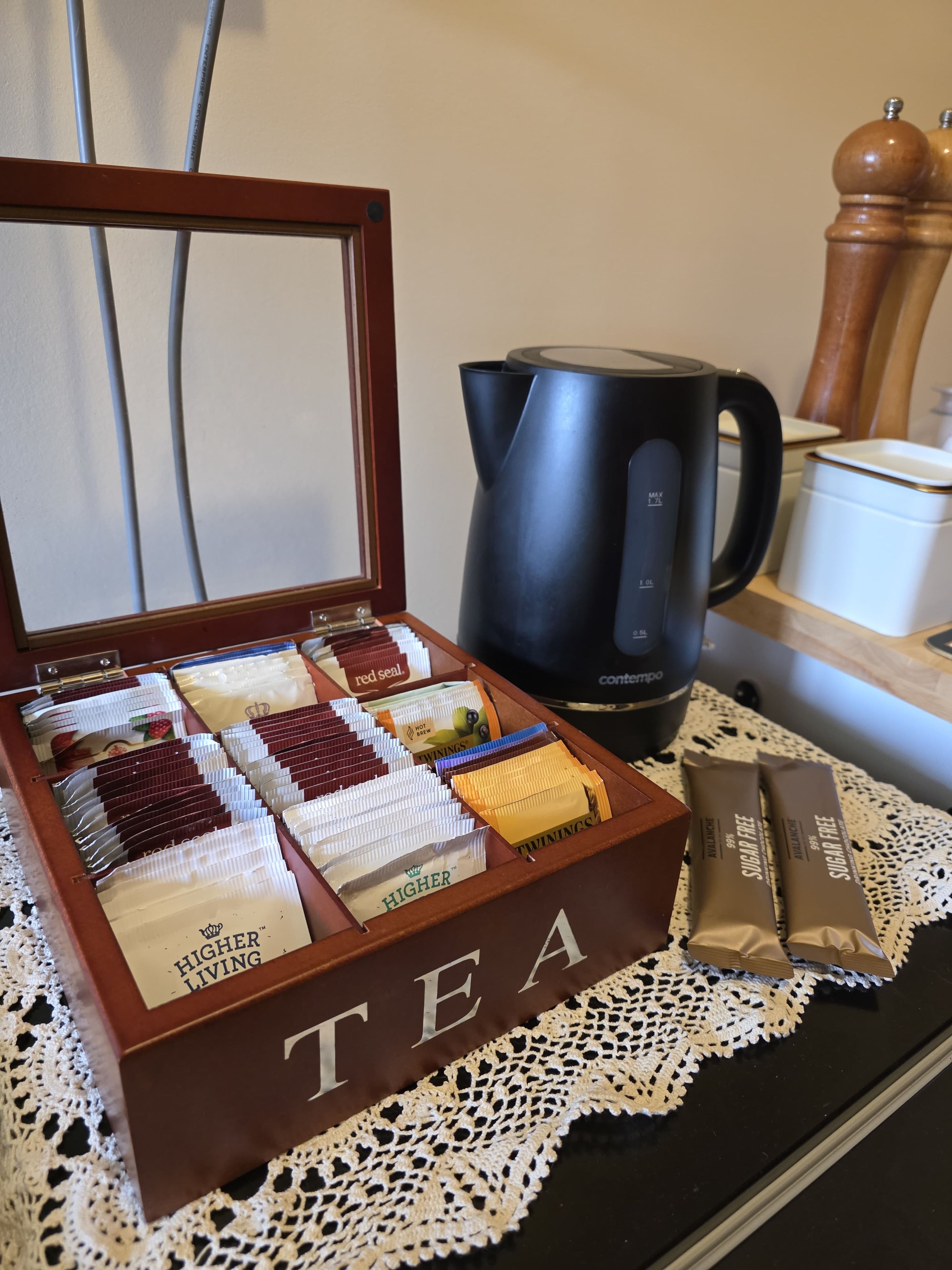 A tea box with compartments containing various tea bags, a black coffee maker, and a sugar-free tea packet on a table with a lace doily.