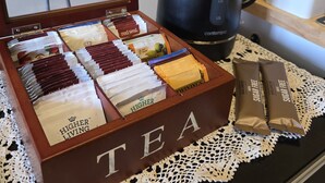Une boîte à thé compartimentée contenant différents sachets de thé, une machine à café noire et un paquet de thé sans sucre, le tout posé sur une table recouverte d’une nappe en dentelle.