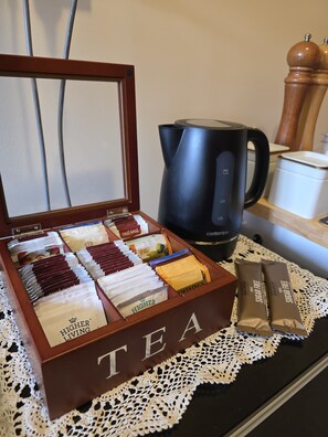 A tea box with compartments containing various tea bags, a black coffee maker, and a sugar-free tea packet on a table with a lace doily. - 🌿 Private Queen Bedroom, Private Bathroom & Garden Retreat in Sunbury (Sunbury)