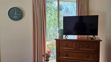 A room with a wooden dresser, a television on top, a clock on the wall, and a window with curtains.