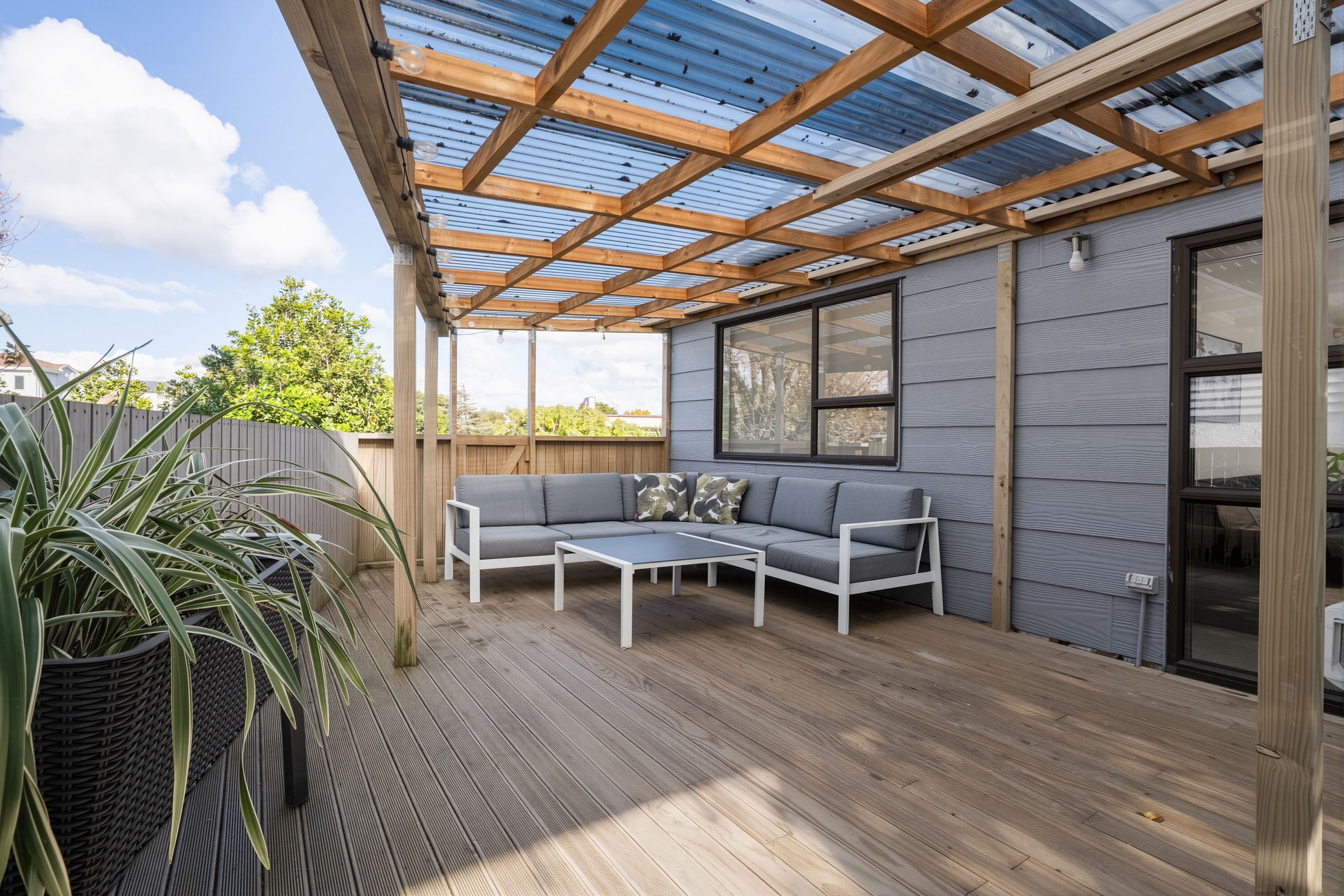 A covered patio with a wooden deck, a grey sofa, and a table.