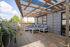A covered patio with a wooden deck, a grey sofa, and a table. - Newly Renovated 4BR 2.5B Home | Sleeps9 | 3Parking (Auckland)