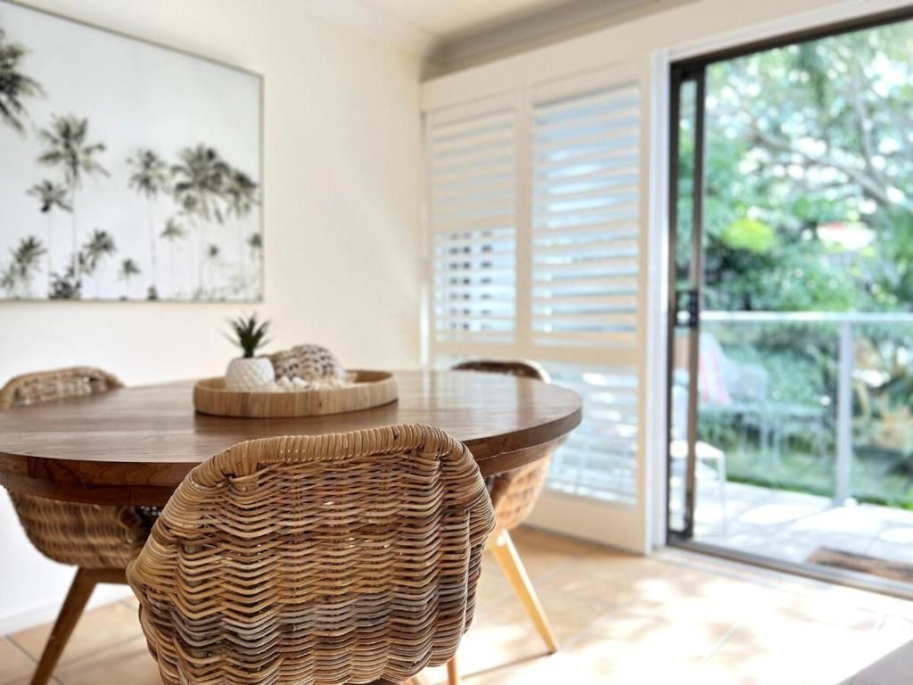 A dining area with a round table, wicker chairs, and a view of the outdoors through large windows.