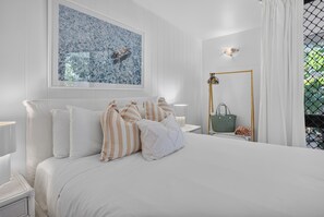 A neatly arranged bedroom with a large bed, a framed artwork above it, and a view of greenery through a window.