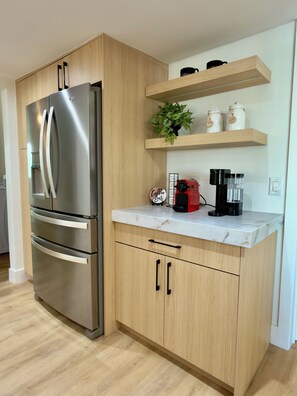 Eine moderne Küche mit einem Edelstahl-Kühlschrank, Holzschränken, einer Marmorarbeitsplatte, einer Kaffeemaschine und einem Regal mit Gläsern und einer Pflanze.