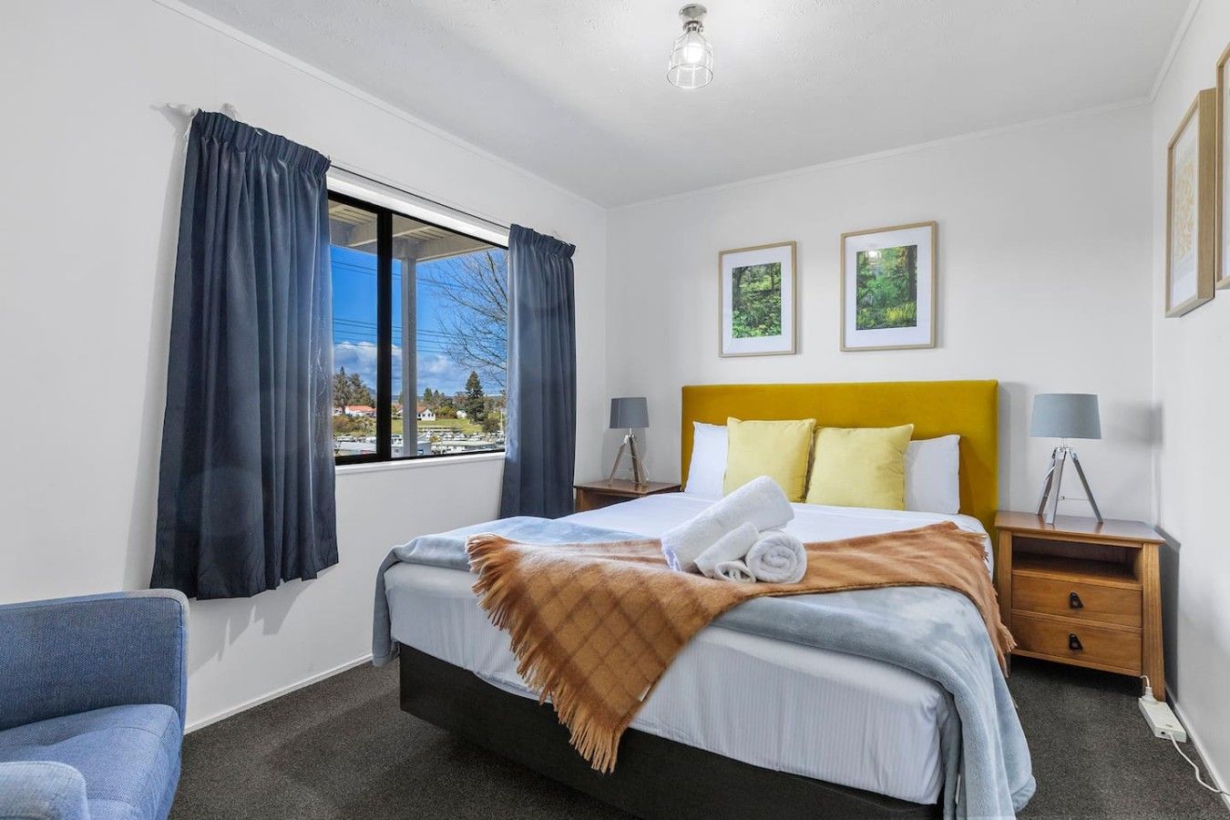 A hotel room with a bed, a bedside table with a lamp, a window with curtains, and two framed pictures on the wall.