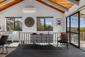 Ein Zimmer mit weißer Decke, einem Holzbalken, einem Fenster mit weißem Rahmen, einem an der Wand befestigten dekorativen Teller, einer weißen Kommode, einem Stuhl und Blick ins Freie.