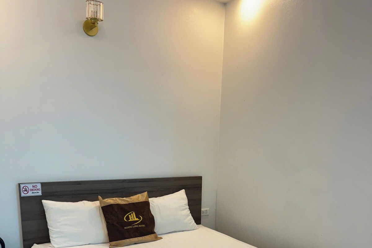 A hotel room with a bed, a nightstand, a wall-mounted light, and a 'No Smoking' sign.