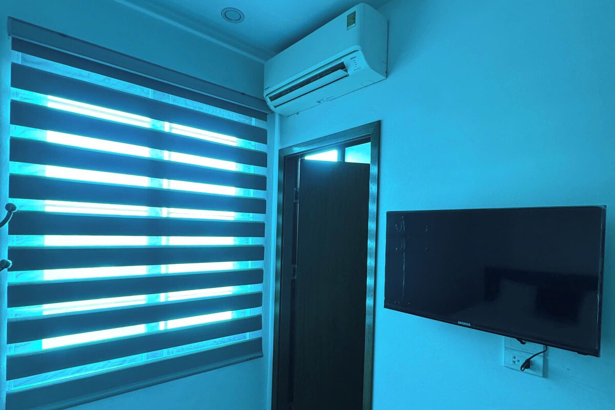 A room with a television mounted on the wall, a window with horizontal blinds, and an air conditioning unit on the ceiling.