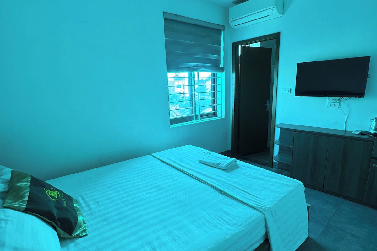 Quarto com uma cama de solteiro, janela com persianas, TV de tela plana fixada na parede e porta que dá para outro cômodo.