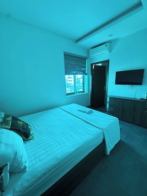 Quarto com uma cama de solteiro, janela com persianas, TV de tela plana fixada na parede e porta que dá para outro cômodo.