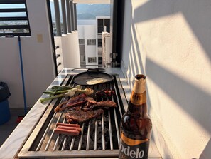 A grill with various meats and vegetables, a bottle of Modelo beer, and a view of buildings in the background. - Acapulco shines! Beautiful apartment. Just 15 minutes from Puerto Márquez and P. Diamante (Acapulco de Juárez)