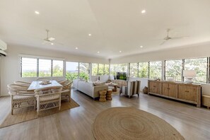 A spacious living area with a dining table, sofas, and a large window offering a view of greenery. - Luxe Sunshine Beach House - 30m to Beach! (Sunshine Beach)