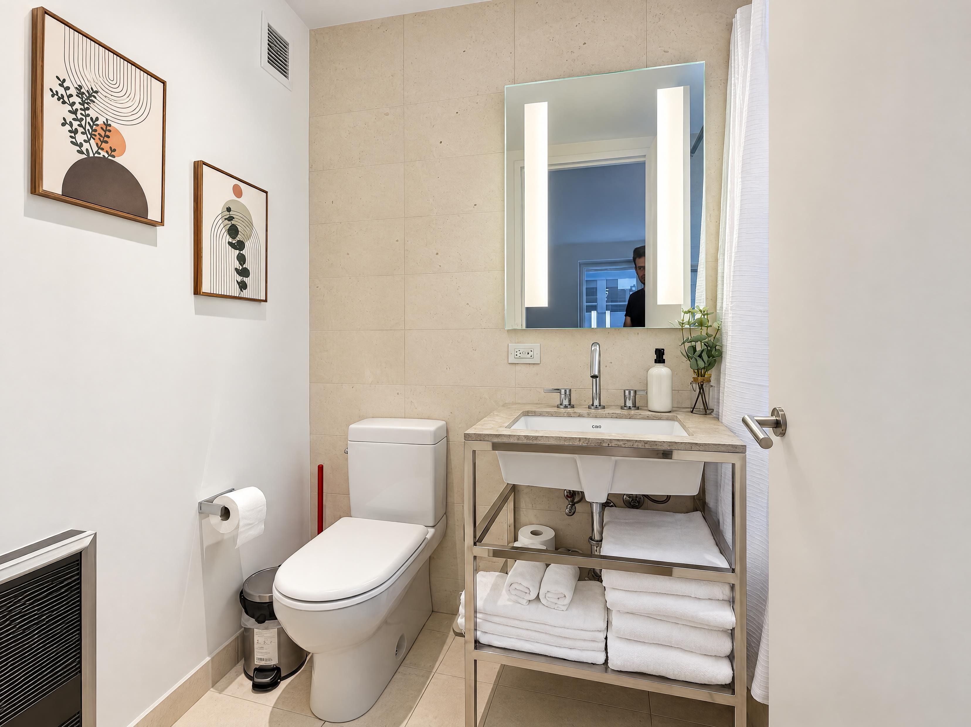 Un baño moderno con inodoro, lavamanos y espejo. Hay toallas y un cesto de basura.