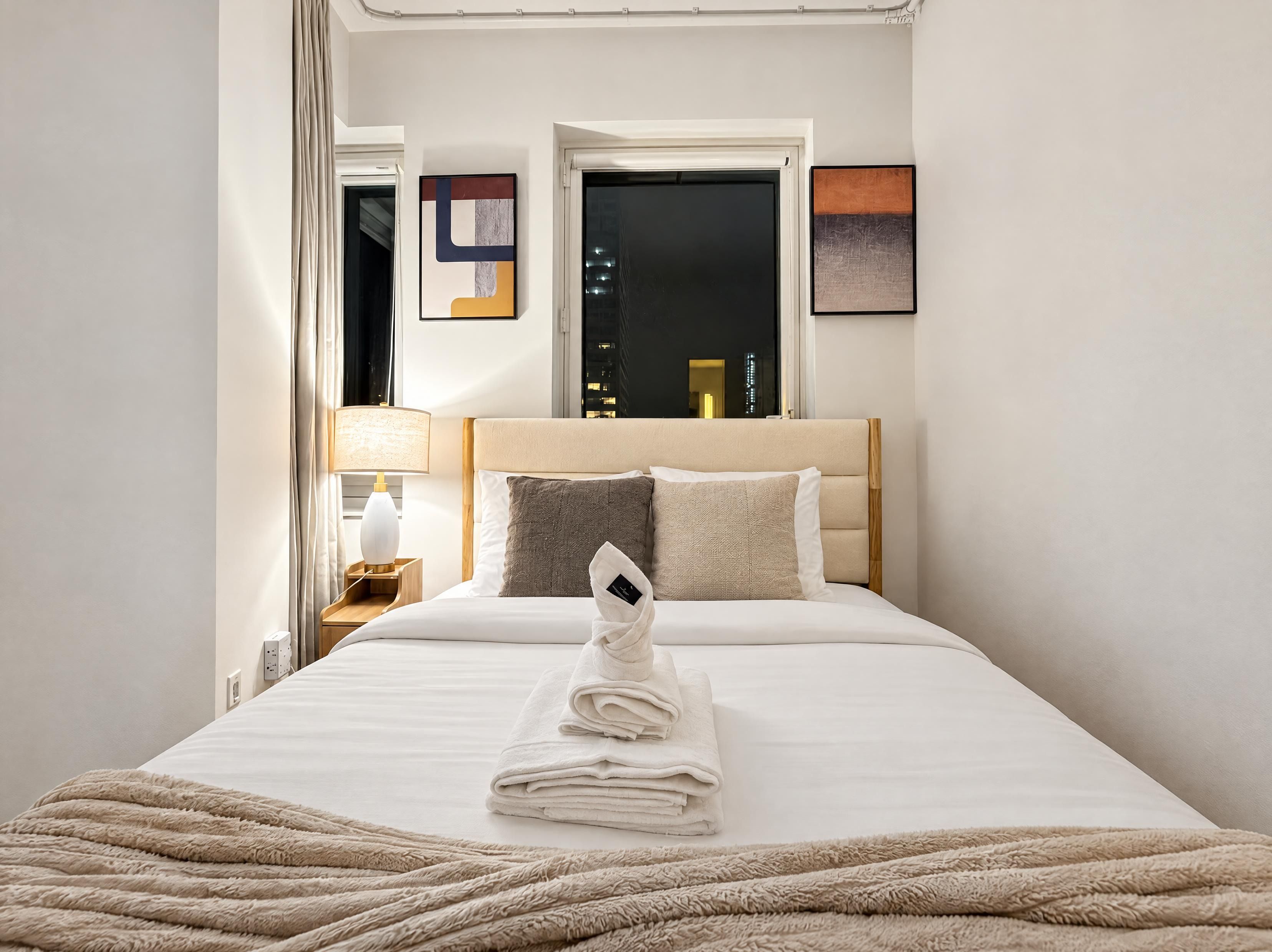 A hotel room with a bed, a nightstand, a lamp, and a window with a city view.
