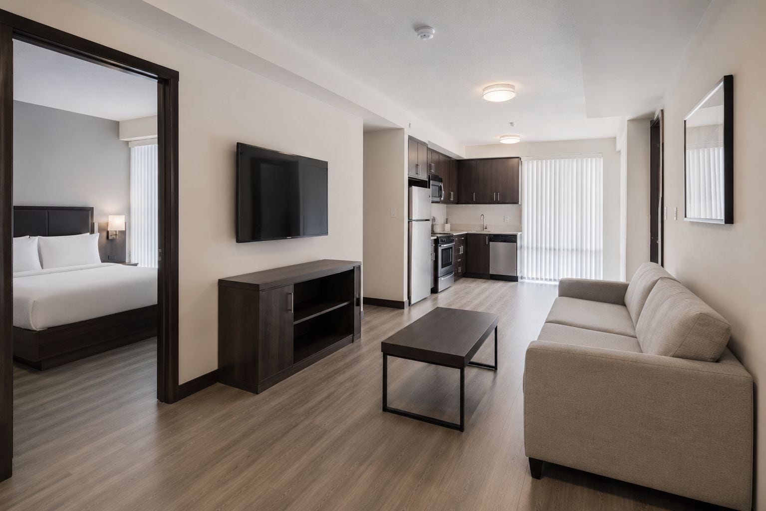 Una habitación de hotel moderna con cama, televisor, mueble bajo de madera oscura, una mesita, un sofá y una kitchenette.