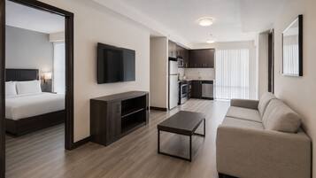 Una habitación de hotel moderna con cama, televisor, mueble bajo de madera oscura, una mesita, un sofá y una kitchenette.