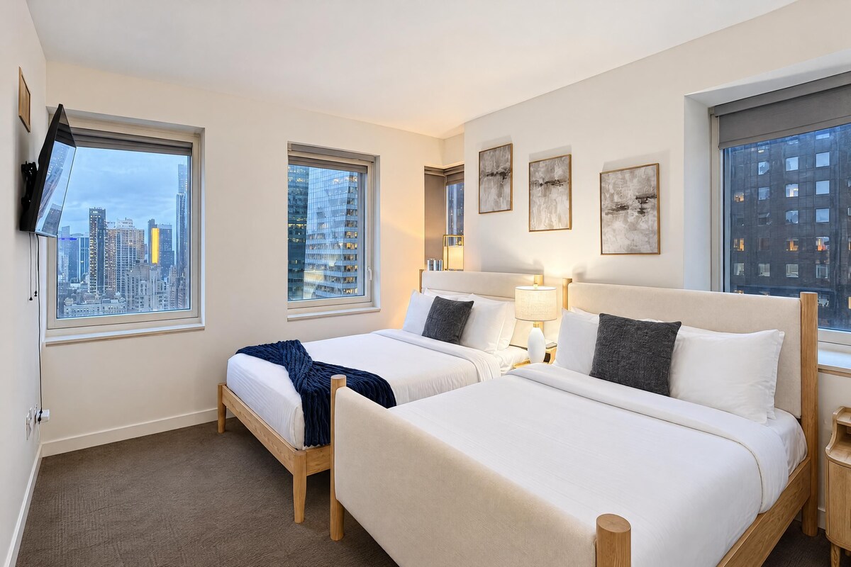A hotel room with two beds, a TV mounted on the wall, and large windows offering a city view.