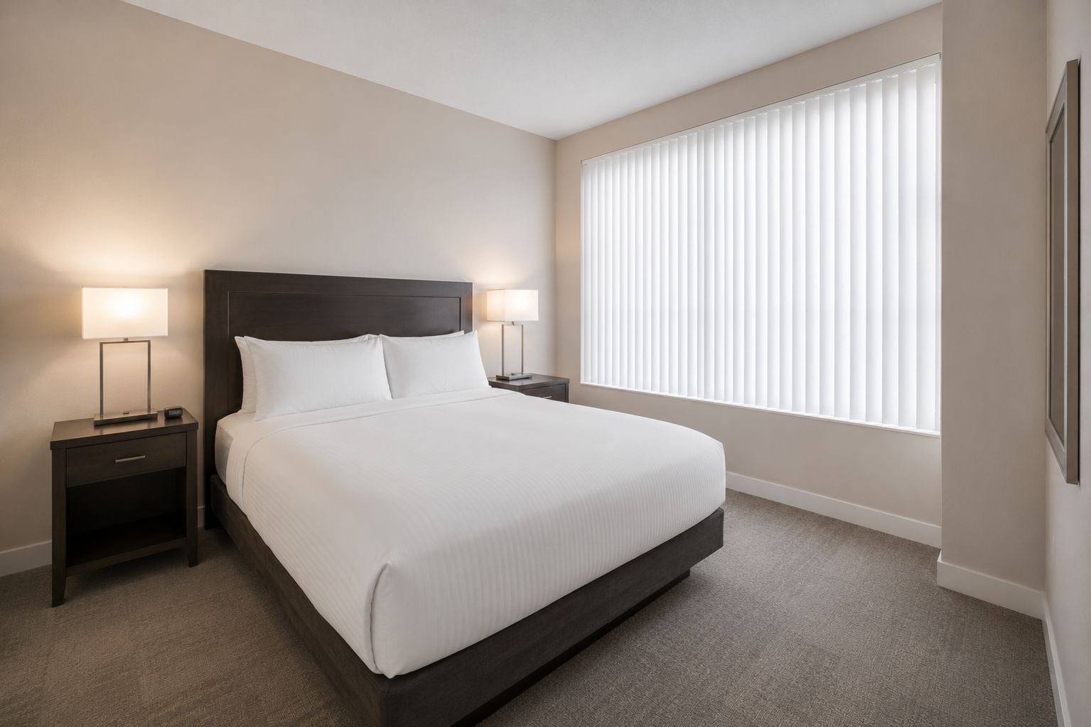 A hotel room with a large bed, two bedside tables with lamps, a window with blinds, and a carpeted floor.