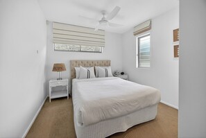 A bedroom with a bed, a nightstand, a lamp, a ceiling fan, and a window. - Sunshine Luxe Penthouse - Wide Ocean Views (Sunshine Beach)