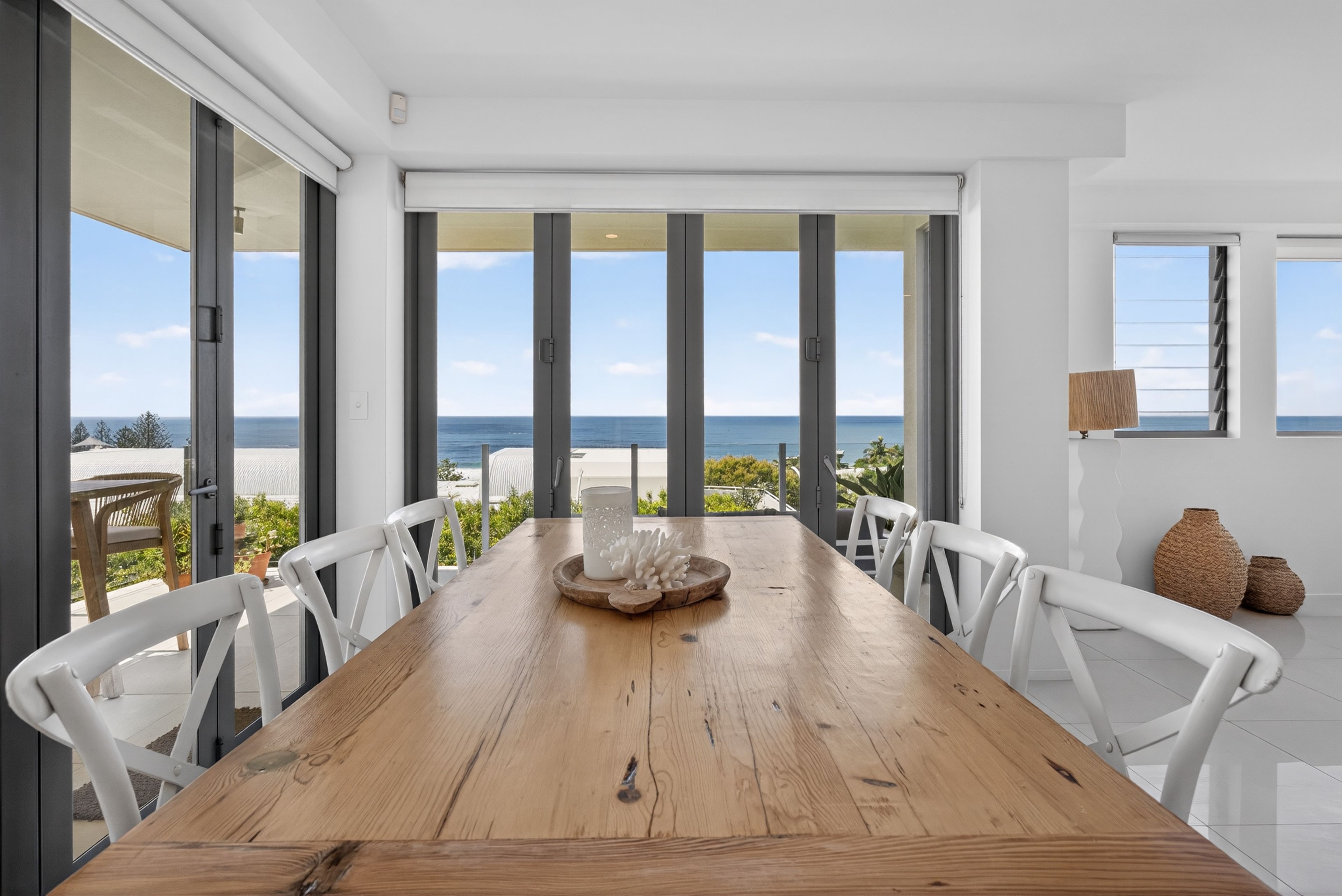 Un comedor con mesa de madera y sillas blancas, ventanas amplias con vista al mar, y una playa visible al exterior.