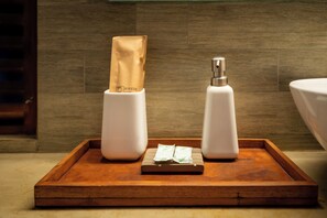 Una bandeja con dos recipientes blancos, uno con una bolsa de papel marrón y el otro con una botella plateada, sobre una superficie de madera en un baño.